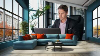 Young businessman wearing black suit, sitting to table with laptop. Office manager working in modern office with glass windows blurred background, glass of water on the table, concept of work Wall mural