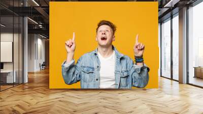 Amazed young man in denim casual clothes keeping mouth open pointing index finger up isolated on yellow orange wall background in studio. People sincere emotions lifestyle concept. Mock up copy space. Wall mural