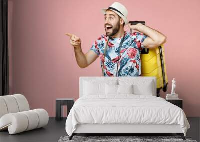 Amazed young traveler tourist man in summer clothes hat photo camera hold suitcase pointing index finger aside isolated on pink background. Passenger traveling on weekend. Air flight journey concept. Wall mural