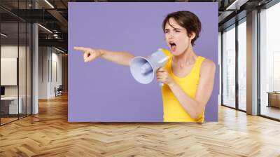 Angry young brunette woman girl in yellow casual tank top posing isolated on pastel violet wall background studio portrait. People lifestyle concept. Screaming in megaphone point index finger aside. Wall mural