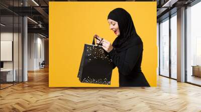 Arabian muslim woman in black hijab hold package bag, purchases after shopping isolated on yellow background studio portrait. Birthday holiday people religious lifestyle concept. Mock up copy space. Wall mural