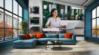 Attractive young modern business woman working with documents and laptop in the kitchen at home Wall mural