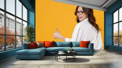 Back rear view of funny young business woman in white shirt glasses isolated on yellow background. Achievement career wealth business concept. Mock up copy space. Showing thumb up, looking camera. Wall mural