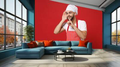 Bearded male chef cook or baker man in striped apron toque chefs hat posing isolated on red background. Cooking food concept. Saying hush be quiet with finger on lips shhh gesture, try to hear you. Wall mural