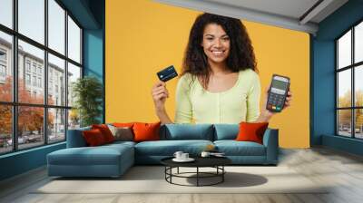 Beautiful african american young brunette woman 20s wears green shirt hold wireless modern bank payment terminal to process acquire credit card payments isolated on yellow background studio portrait Wall mural