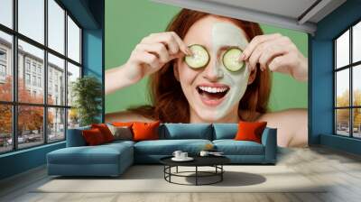 Beautiful fun half naked topless redhead hair woman 20s apply clay facial mask cover two eyes with cucumber isolated on pastel green color background Skin care healthcare cosmetic procedures concept Wall mural