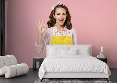 Blinking young brunette woman housewife 20s in yellow apron showing OK gesture looking camera while doing housework isolated on pastel pink colour background studio portrait. Housekeeping concept. Wall mural