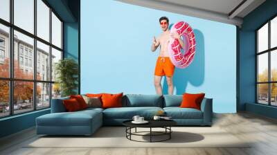 Blinking young man in orange shorts glasses hold inflatable ring isolated on blue background studio. People summer vacation rest lifestyle concept. Mock up copy space. Pointing index finger on camera. Wall mural