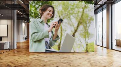 Bottom view young student freelancer woman 20s in green jacket jeans sit on bench in spring park outdoors rest use laptop pc computer talk speak by mobile cell phone. People urban lifestyle concept Wall mural