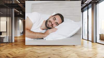 Calm young handsome bearded man lying in bed with white sheet pillow blanket in bedroom at home. Smiling beauty male spending time in room. Rest relax good mood lifestyle concept. Mock up copy space. Wall mural