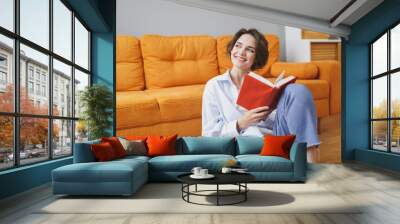 Charming young woman girl in white casual clothes sitting on floor near couch spending time in living room at home. Rest relax good mood leisure lifestyle concept. Mock up copy space. Reading book. Wall mural