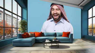 Cheerful bearded young arabian muslim man in keffiyeh kafiya ring igal agal casual clothes posing isolated on pastel blue background. People religious lifestyle concept. Mock up copy space. Blinking. Wall mural