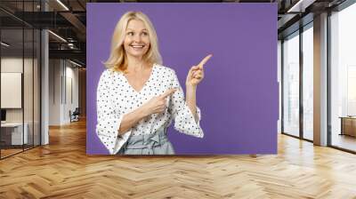 Cheerful elderly gray-haired blonde woman lady 40s 50s years old wearing white dotted blouse standing point index fingers aside up on mock up copy space isolated on violet background studio portrait. Wall mural