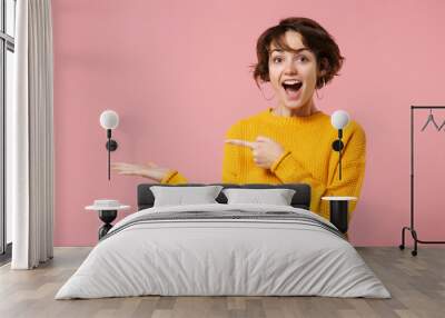 Cheerful excited young brunette woman girl in yellow sweater posing isolated on pastel pink background studio portrait. People lifestyle concept. Mock up copy space. Pointing index finger, hand aside. Wall mural