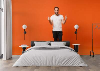 Cheerful excited young man in casual white t-shirt posing isolated on bright orange wall background studio portrait. People sincere emotions lifestyle concept. Mock up copy space. Showing thumbs up. Wall mural