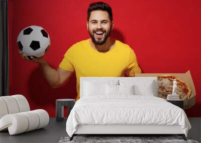 Cheerful fun young bearded man football fan in yellow t-shirt cheer up support favorite team hold soccer ball italian pizza in cardboard flatbox isolated on plain dark red background studio portrait. Wall mural