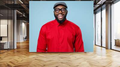 Cheerful laughing handsome young bearded african american man 20s wearing casual red shirt cap eyeglasses standing and looking camera isolated on pastel blue color wall background studio portrait. Wall mural