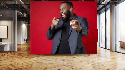 Cheerful laughing successful young african american business man 20s wearing classic jacket suit standing pointing index fingers on camera isolated on bright red color background studio portrait. Wall mural