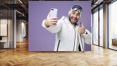 Cheerful skier man in white jacket ski goggles mask doing selfie shot on mobile phone showing thumb up spend weekend winter in mountains isolated on purple background. People lifestyle hobby concept. Wall mural