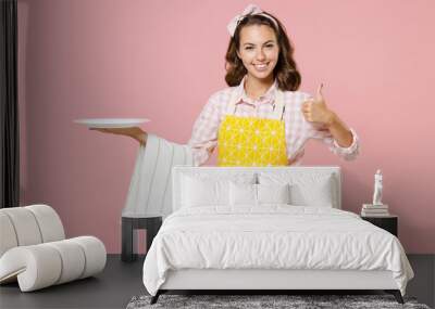 Cheerful smiling young woman housewife 20s in yellow apron hold empty plate dishcloth towel showing thumb up doing housework isolated on pastel pink background studio portrait. Housekeeping concept. Wall mural