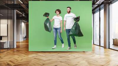 Cheerful two young friends couple in white volunteer t-shirt isolated on pastel green background. Voluntary free work assistance help charity grace teamwork concept. Holding trash bags, looking aside. Wall mural