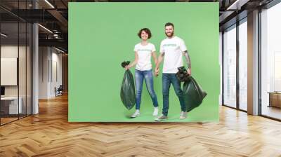 Cheerful two young friends couple in white volunteer t-shirt isolated on pastel green wall background. Voluntary free work assistance help charity grace teamwork concept. Holding hands, trash bags. Wall mural