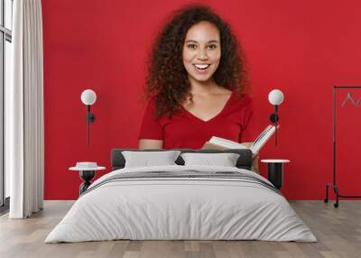 Cheerful young african american woman girl in casual t-shirt posing isolated on bright red wall background studio portrait. People sincere emotions lifestyle concept. Mock up copy space. Reading book. Wall mural