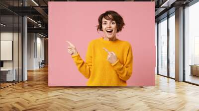 Cheerful young brunette woman girl in yellow sweater posing isolated on pastel pink background studio portrait. People sincere emotions lifestyle concept. Mock up copy space. Point index finger aside. Wall mural