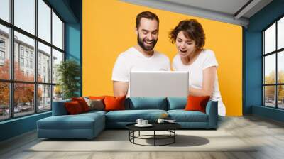 Cheerful young couple friends bearded guy girl in white blank empty t-shirts isolated on yellow orange wall background. People lifestyle concept. Mock up copy space. Working on laptop pc computer. Wall mural