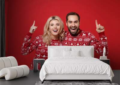 Cheerful young couple guy girl in Christmas sweaters posing isolated on red background studio portrait. Happy New Year 2020 celebration party concept. Mock up copy space. Pointing index fingers up. Wall mural