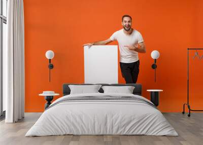 Cheerful young man in casual white t-shirt posing isolated on orange wall background in studio. People lifestyle concept. Mock up copy space. Pointing index finger on big white empty blank billboard. Wall mural