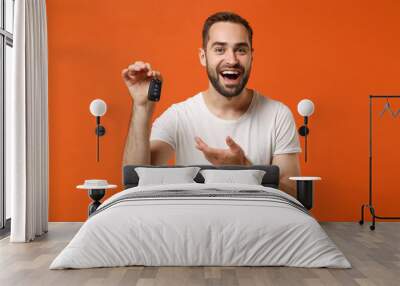 Cheerful young man in casual white t-shirt posing isolated on orange wall background studio portrait. People sincere emotions lifestyle concept. Mock up copy space. Holding, pointing hand on car keys. Wall mural