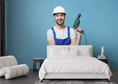 Cheerful young man in coveralls protective helmet hardhat hold electric drill isolated on pastel blue background in studio. Instruments accessories for renovation apartment room. Repair home concept. Wall mural