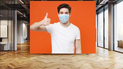 Cheerful young man wearing casual white t-shirt sterile face mask to safe from coronavirus virus covid-19 during pandemic quarantine showing thumb up isolated on orange background studio portrait. Wall mural