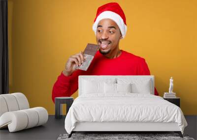 Cheerful young Santa african american man wearing red sweater Christmas hat biting eating chocolate bar isolated on yellow background studio portrait. Happy New Year celebration merry holiday concept. Wall mural