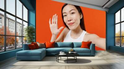 Close up charming smiling young woman of Asian ethnicity 20s years old in white tank top doing selfie shot pov on mobile phone meet greet with hand isolated on plain orange background studio portrait. Wall mural