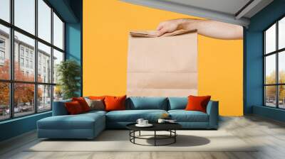 Close up cropped male hold brown empty blank paper bag for takeaway isolated on yellow background. Food delivery service from restaurant shop to home. Epidemic pandemic coronavirus 2019-ncov covid-19. Wall mural