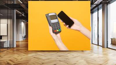 Close up cropped photo female holding in hands wireless modern bank payment terminal to process and acquire credit card payments mobile phone isolated on yellow background. Copy space for advertising. Wall mural