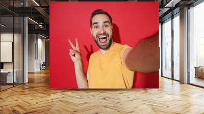 Close up of cheerful young bearded man guy in casual yellow t-shirt isolated on red background. People lifestyle concept. Mock up copy space. Doing selfie shot on mobile phone, showing victory sign. Wall mural