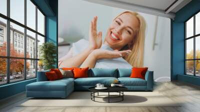 Close up young amazed smiling happy woman covered by napkin spread hands sit at dental office chair indoor light modern cabinet wait stomatologist for oral procedure Healthcare caries enamel treatment Wall mural