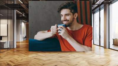 Close up young man wears red t-shirt hold cup drink hot tea look aside sit on blue sofa stay at home hotel flat rest relax spend free spare time in living room indoors grey wall People lounge concept Wall mural