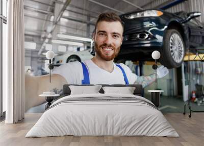 Close up young professional technician mechanic man in blue overalls t-shirt do selfie shot pov mobile phone stand near car lift point hand on automobile work in vehicle repair shop workshop indoors Wall mural