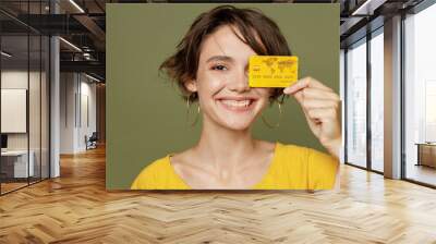 Close up young smiling satisfied fun happy woman she 20s wear yellow t-shirt holding in hand cover eye with mock up of credit bank card isolated on plain olive green khaki background studio portrait Wall mural