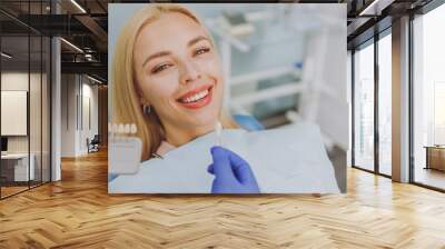 Close up young smiling woman choosing veneers enamel color palette with stomatologist compare teeth shade sit at dentist office chair indoor light modern cabinet Healthcare enamel whitening treatment. Wall mural