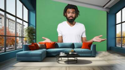 Confused puzzled young african american man guy with dreadlocks 20s wearing white casual t-shirt posing spreading hands looking camera isolated on green color wall background studio portrait. Wall mural