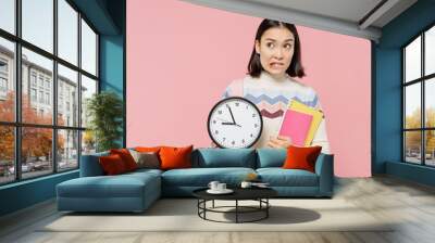 Confused sad unhappy teen student girl of Asian ethnicity wearing sweater backpack hold books clock isolated on pastel plain light pink background Education in high school university college concept. Wall mural