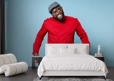 Confused smiling young bearded african american man 20s wearing casual red shirt eyeglasses cap has no money showing empty pockets looking camera isolated on pastel blue background studio portrait. Wall mural