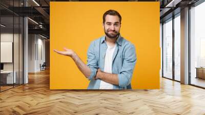 Confused young bearded man in casual blue shirt posing isolated on yellow orange wall background, studio portrait. People sincere emotions lifestyle concept. Mock up copy space. Pointing hand aside. Wall mural