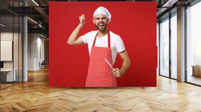 Crazy young bearded male chef cook or baker man in striped apron toque chefs hat posing isolated on red background. Cooking food concept. Mock up copy space. Holding cutlery fork and knife, screaming. Wall mural