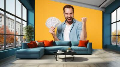 Crazy young man in blue shirt posing isolated on yellow orange background. People lifestyle concept. Mock up copy space. Holding fan of cash money in dollar banknotes, doing winner gesture, screaming. Wall mural